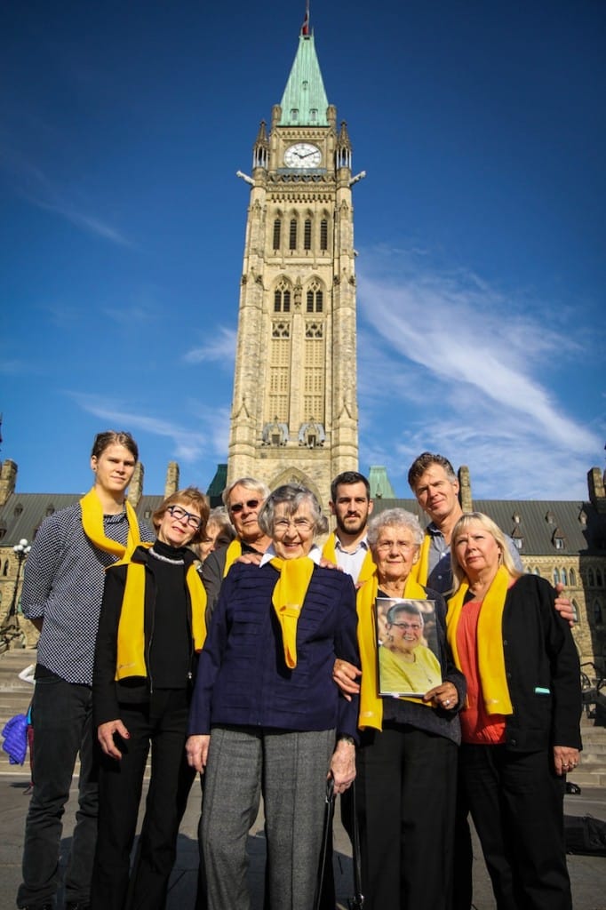 Kay Carter and Gloria Taylor's families stand smiling in front of Parliament Hill while holding up a portrait of Gloria Taylor. They are wearing yellow to commemorate Gloria. 