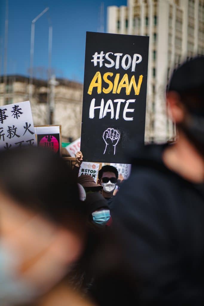 Person among crowd in sunglasses and a white face mask holding a black and white sign that says "#Stop Asian Hate" with a drawn fist. Two blurred people are at the foreground of image, one in black shirt. 